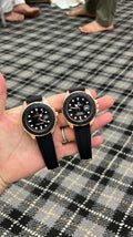 Two wristwatches with black straps and black dials are held in a person's hand over a black-and-white plaid carpet. The watches have gold-toned cases and bezels, with white hour markers and date windows.